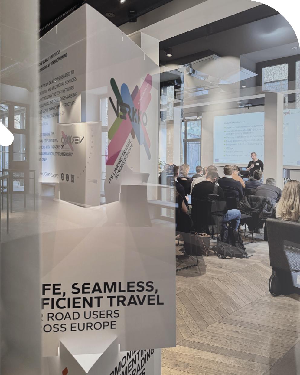 Teilnehmende sitzen in einem Konferenzraum und verfolgen eine Präsentation beim Steering Committee Meeting des EU-Projekts VERKKO in Berlin. Im Vordergrund steht ein Banner mit dem VERKKO-Logo und dem Schriftzug zu vernetztem Verkehr in Europa.