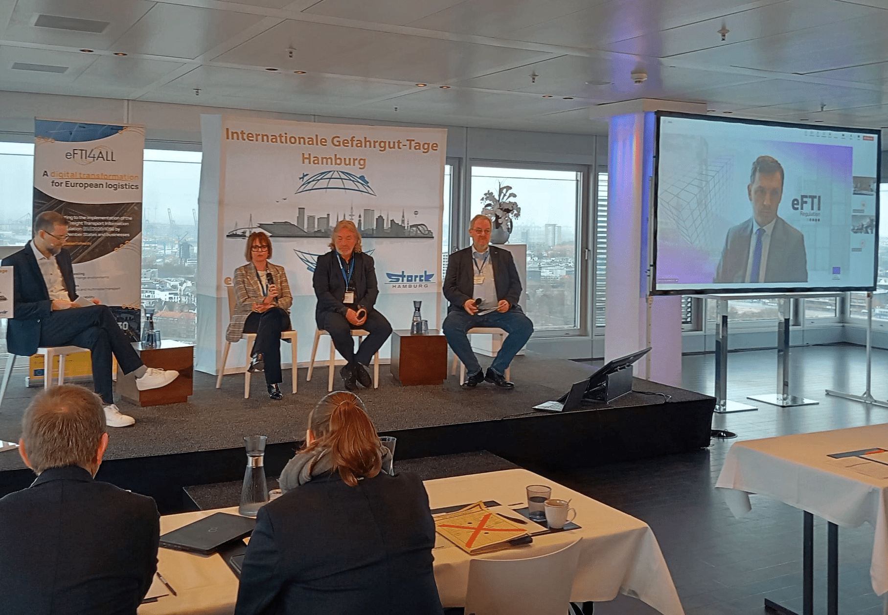Four speakers are seated on a small stage in a conference room, holding a panel discussion in front of an audience. Behind them are banners reading “Internationale Gefahrgut-Tage Hamburg” and “eFTI4ALL”. On the right, a large screen shows another participant joining via video call with the word “eFTI” displayed. Through the large windows in the background, the skyline of Hamburg is visible.
